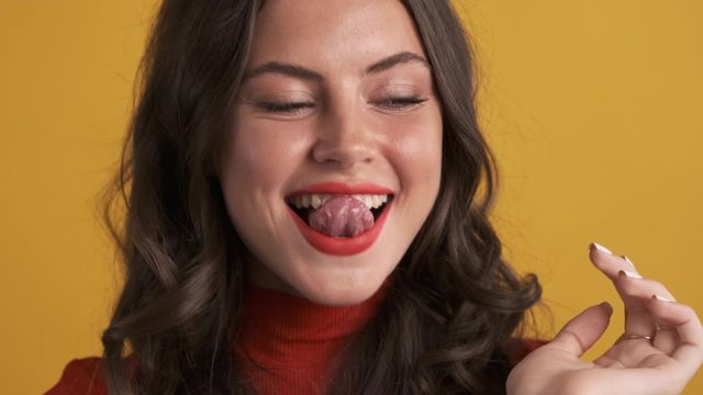 Portrait of attractive brunette girl happily licking teeth on camera over yellow background