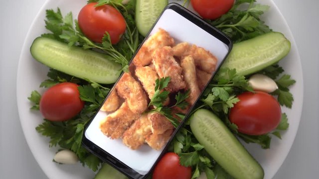 Plate with salad and cellphone with crispy fried cod fish fillet on screen. Food blog concept. Top view of mobile phone with food footage on screen rotating.