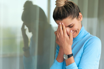 Horizontal portrait of beautiful young woman smiling and posing next the window in office wearing blue turtleneck, with fingers touching her forehead. People and business concept