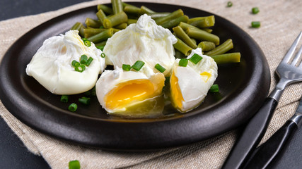 Close-up poached eggs with asparagus beans and chopped onion on the black plate. Concept of health and fast everyday breakfast. Grey background, spoon, fork and rough cloth
