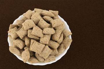 Dry breakfast cereal with chocolate filling. Bowl with chocolate pads on blurred chocolate color felt background. Selective focus