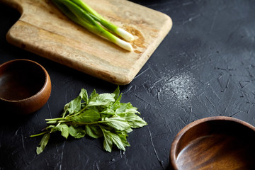 Cooking dinner. Cutting board and herb ingredients. Fresh mint leaves, scallion (green onion), empty natural wooden salad bowls on stone table, black background