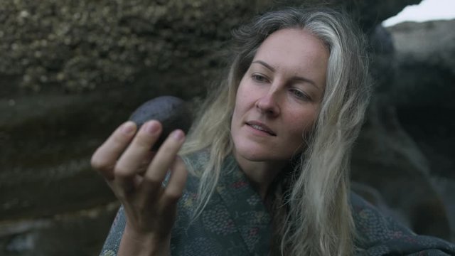 Young woman with strand of natural gray hair holds in the palm a stone and stares at him by the ocean