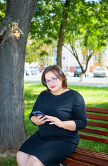 Beautiful chubby girl in a black strict dress sits on a bench in the park with a phone in his hand