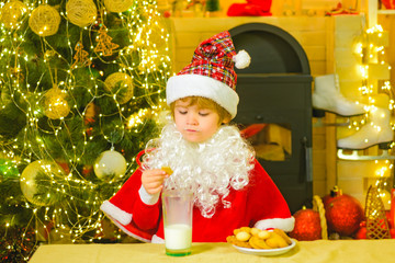 Little Santa Claus kid with beard and mustache. Santa - funny child picking cookie. Portrait of little Santa child holding chocolate cookie and glass of milk.