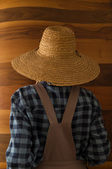 the woman wearing county style clothes and wicker hat stands in front of wooden wall.