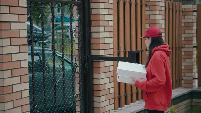 A pizza delivery man in red uniform brought two boxes of pizza. Happy girl phoning an answering machine to a customer. A call to the intercom to open the gate.