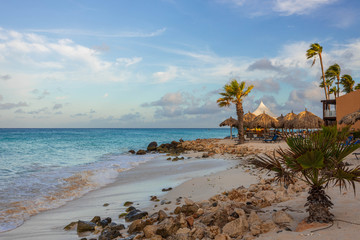 Gorgeous view of Aruba coast line landscape. Turquoise water of Atlantic ocean, white sand beach with palm trees and sun umbrellas on blue sky with white clouds background.