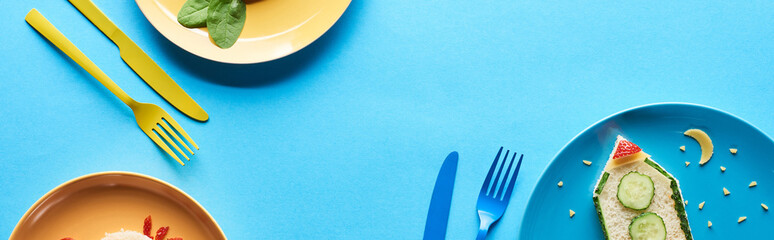 top view of plates with fancy childrens breakfast near cutlery on blue background, panoramic shot