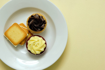 Creamy mouth-watering cupcakes and biscuits served on a white plate against a yellow background - top view, food design template with copy space for text on the side.