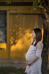 Beautiful asian pregnant woman smiling and standing under a tree make hopefully face with wooden yellow hose wall with shadow of tree.