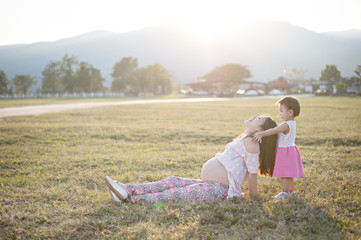 baby girl  hugging asian beautiful pregnant woman on grass field in evening light with sunset and mountain background, Good health and relax concept, family relationship.
