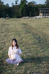 Beautiful asian pregnant woman smiling and siting making a heart sign on grass field with sunset and mountain background.