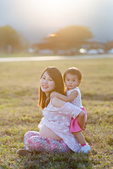 baby girl  hugging asian beautiful pregnant woman on grass field in evening light with sunset and mountain background, Good health and relax concept, family relationship.