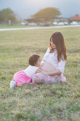 Naklejka na ściany i meble baby girl hugging asian beautiful pregnant woman on grass field in evening light with sunset and mountain background, Good health and relax concept, family relationship.