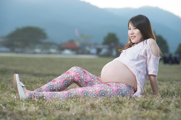 Beautiful asian pregnant woman smiling and siting making a heart sign on grass field with sunset and mountain background.