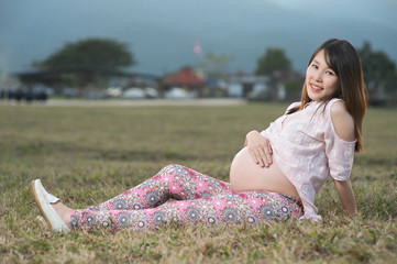 Beautiful asian pregnant woman smiling and siting making a heart sign on grass field with sunset and mountain background.