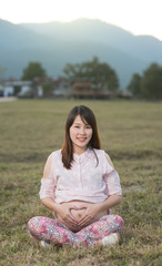 Beautiful asian pregnant woman smiling and siting making a heart sign on grass field with sunset and mountain background.