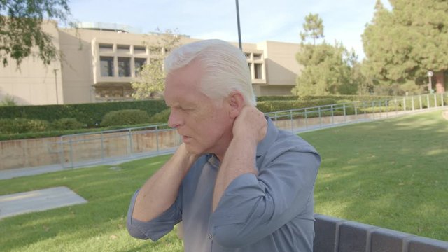 Closeup Portrait, Senior Man Experiencing Excruciating Back Neck Pain With Limited Range Of Motion, Isolated Park Bench Outside Outdoors Background