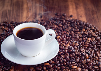 Composition of roasted coffee beans pilled round white cup full of aromatic espresso, close up view. Texture of brown wooden table. Selective soft focus. Blurred background