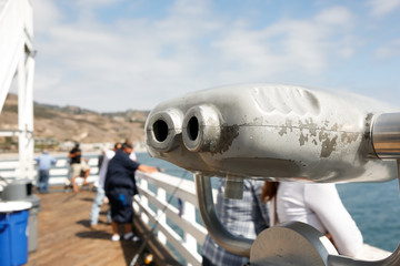 A closeup view of pier binoculars, featuring people in the background fishing and enjoying the view.