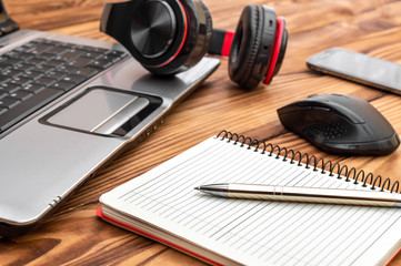 Wooden desk with blank notepad, laptop, smartphone, computer mouse and headphones.