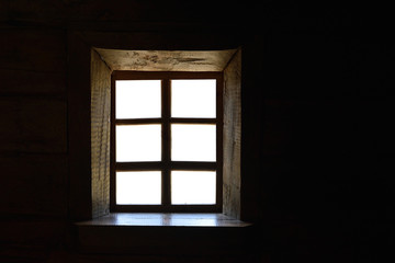 Empty vintage wooden window with white background on a dark wooden wall of an old log house. View from inside darkness