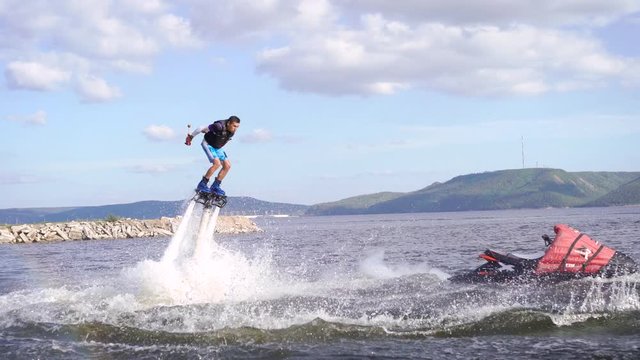 man is flying on flyboard on lake or sea in day, hydrocycle is floating near, water flows is lifting human