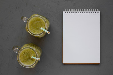 Homemade green cucumber apple smoothie in glass jars, blank notepad on a gray surface, top view. Flat lay, overhead, from above. Copy space.