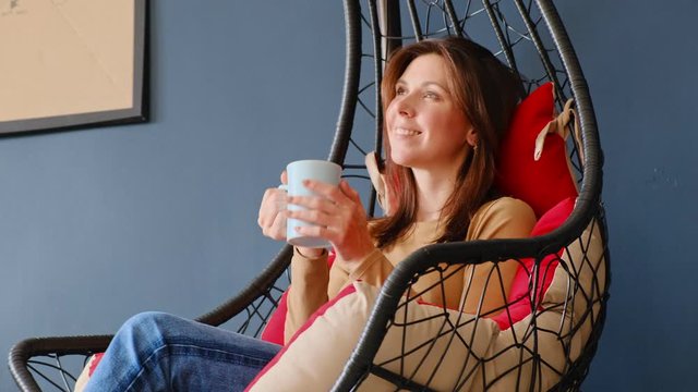 Young attractive girl with cup of tea sitting in hanging chair cocoon and relax.  Blue wall interior design. 4K slow motion video.