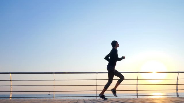 Athletic young girl jogging in the morning by sea wooden embankment. Silhouette of girl in sports costume. Beautiful sunlight. Healthy lifestyle concept.