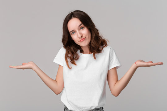 Pretty brunette woman in a white tee shrugging her shoulders beacause she is indignant  standing isolated over grey background. Copy space of advertising.
