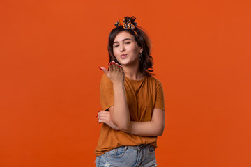 Pretty brunette woman in a t-shirt and beautiful headband sends air kisses standing isolated over orange background. Concept of love