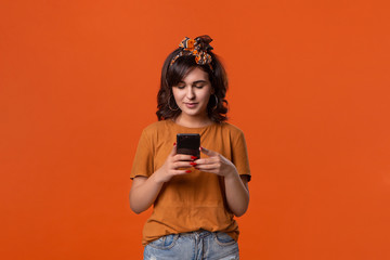 Pretty brunette woman in a t-shirt and beautiful headband checking smartphone for notifications isolated over orange background. Daily communication