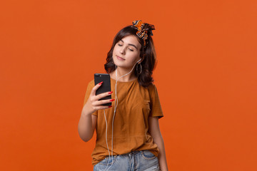 Pretty brunette woman in a t-shirt and beautiful headband making selfie with spartphone standing isolated over orange background. Mobile photography