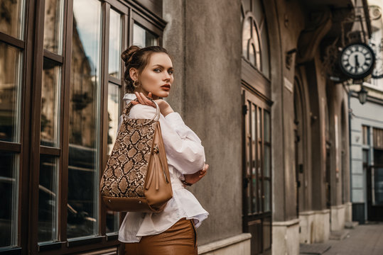 Outdoor fashion portrait of elegant, luxury woman wearing trendy white shirt, holding stylish beige, brown reptile, snakeskin, python print bag, handbag, posing in street. Copy, empty space for text 