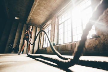 Fitness girl exercising with battle ropes at gym. Woman training doing battling rope workout...