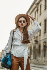 Obraz premium Outdoor fashion portrait of elegant, luxury woman wearing beige hat, black sunglasses, trendy white shirt, leather trousers, with denim patchwork sack bag, posing in street of European city 