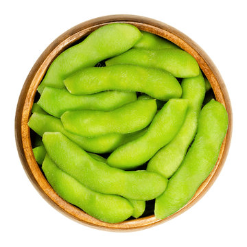 Edamame, green soybeans in the pod, in wooden bowl. Unripe soya beans, also Maodou. Glycine max, a legume, edible after cooking and rich protein source. Closeup, on white background, macro food photo.