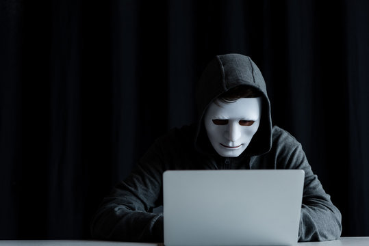 anonymous internet troll in mask typing on laptop keyboard on black