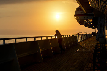 Lone traveller stands on deck of cruise ship watching the beautiful golden sunset as the ship moves across the ocean.