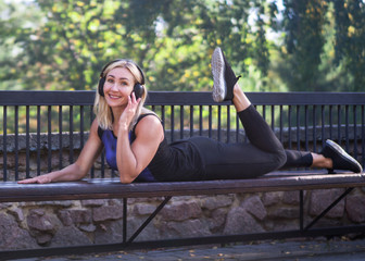 Funny fitness - blond grownup woman take a rest during outdoor workout and listen to the music lying on the bench in urban park, selective focus