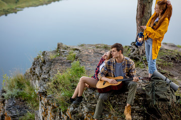 Couple with a guitar resting on a rocky mountain, girl with retro lantern standing behind them and...