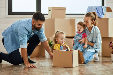Fototapeta na wymiar Caucasian family, man, woman and two girls play, smile with moving box. Mother hugs youngest daughter, father sat eldest daughter into box and move. In background boxes