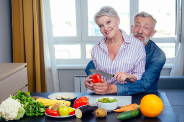 Happy senior couple preparing healthy vegetarian breakfast with fruits and vegetables in day light window background