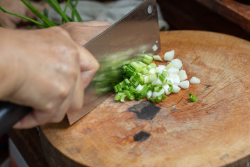 Closeup hands holding knife chopping, cutting green onion, scalliion. Sliced with knife on wooden cutting board. Chopped fresh spring onion..Cooking vegetables in kitchen. Ingredients for Thai foods.