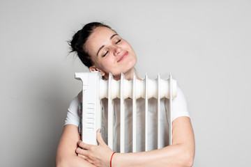 Pleased, young woman hugs an electric radiator and enjoys the warmth, on a white background.