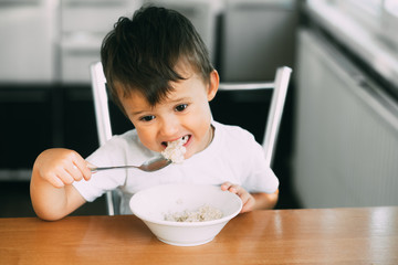 A little boy in the kitchen eating oatmeal from a white plate