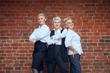 Three adult caucasian blondes businesswoman in office suits pose near brown brick wall. Two of them arms crossed, one of them keep hand in pocket