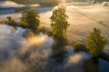 Autumn landscape in morning sunlight from above. Bright sunny misty morning on riverside in september. Vivid sunbeams through fog and trees on river shore